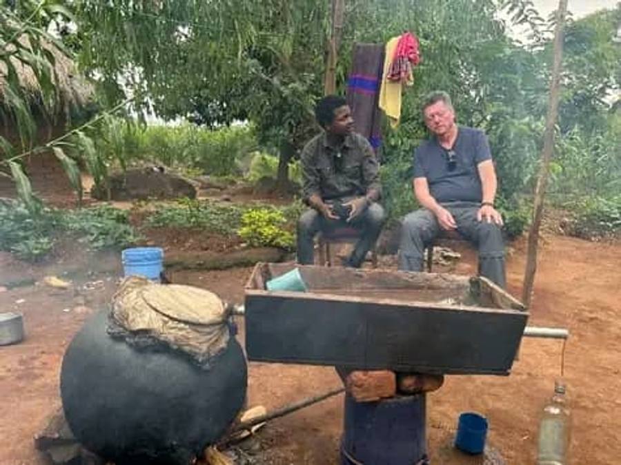 Two men sit beside a large drum still over a fire in a village yard, where kachasu is distilled.