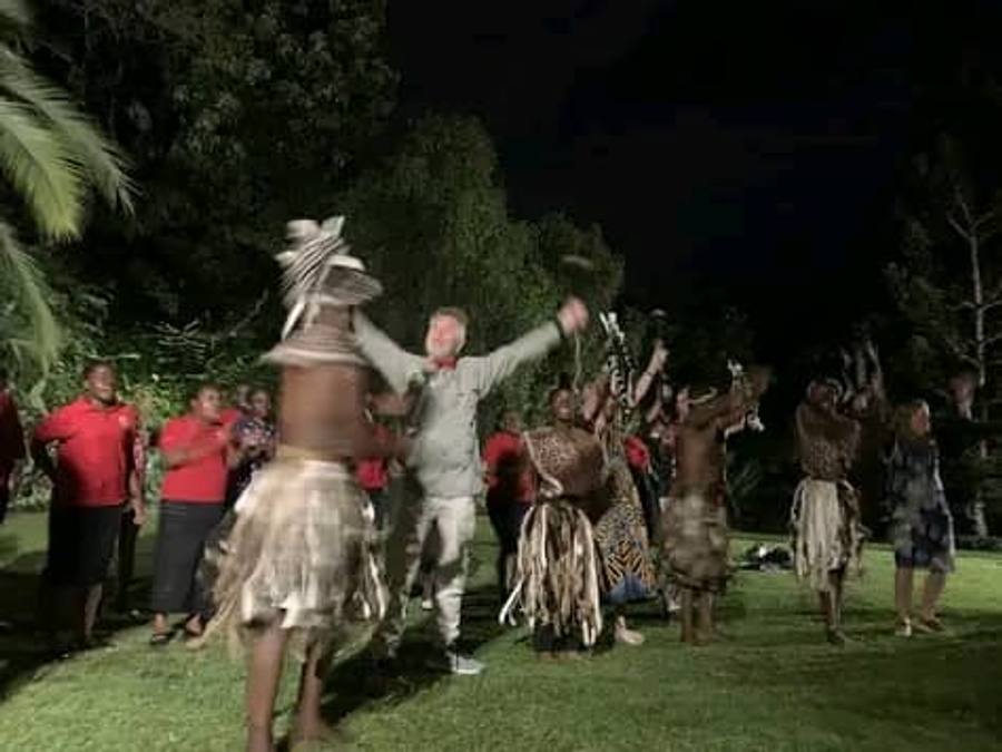 Nyau dancers in raffia skirts and animal-skin costumes perform on floodlit grass at night, a visitor dancing among them and a crowd watching.