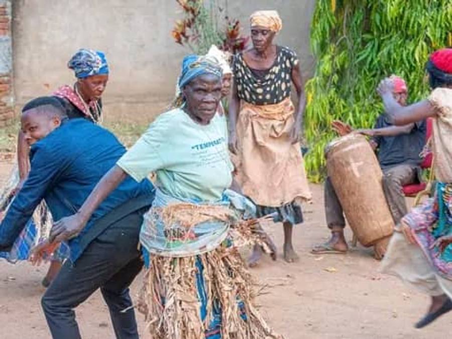 Women of the Theba cultural group dancing in a village yard in grass skirts and headscarves, with a drummer and a bent-forward dancer beside them.